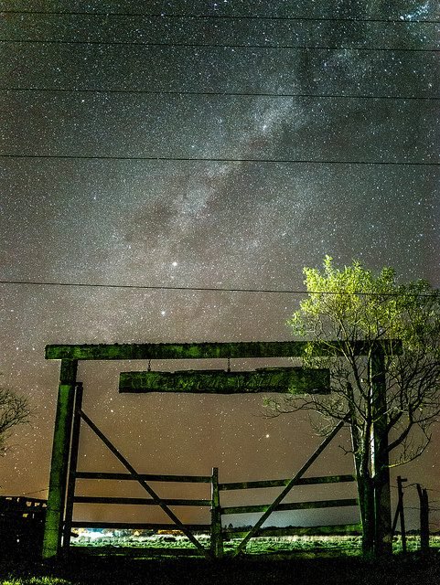La Vía Láctea sobre un portón de madera de campo y un árbol solitario, de noche.