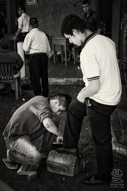 Blanco y negro: un lustrabotas trabajando en el zapato de un cliente en una plaza.