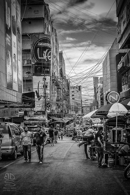 Blanco y negro: una calle comercial llena de gente, con carteles y cables eléctricos enredados.
