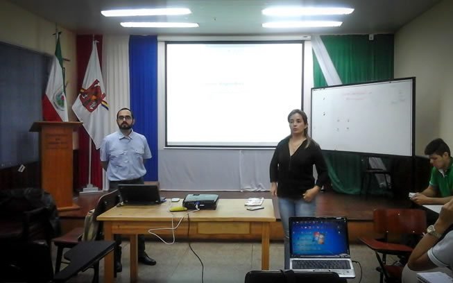 Una capacitación en un aula: un presentador junto a la pantalla del proyector y asistentes con laptops.